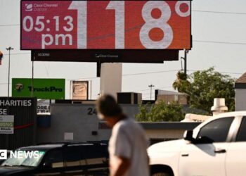 Man walks through road and looks up at sign that says 118 degrees and 05:15 pm