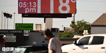 Man walks through road and looks up at sign that says 118 degrees and 05:15 pm