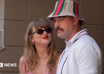 Taylor Swift, wearing a red gingham top, looking up at Travis Kelce, wearing a striped Gucci bucket hat