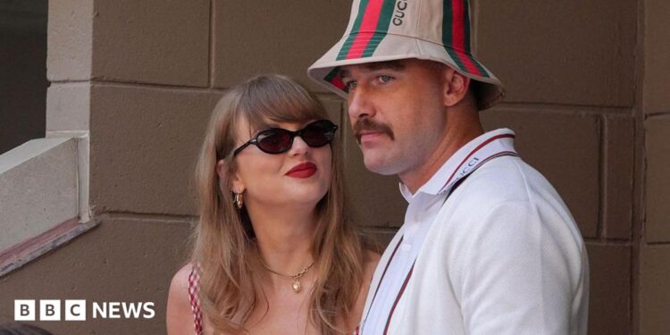Taylor Swift, wearing a red gingham top, looking up at Travis Kelce, wearing a striped Gucci bucket hat