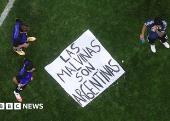 A banner saying "Las Malvinas Son Argentinas" is on the ground of a pitch with three players standing around it