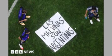 A banner saying "Las Malvinas Son Argentinas" is on the ground of a pitch with three players standing around it
