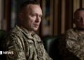 A man sits wearing a military uniform while another uniformed man looks on in the background