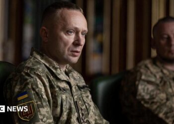A man sits wearing a military uniform while another uniformed man looks on in the background