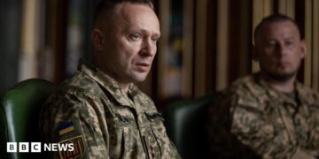 A man sits wearing a military uniform while another uniformed man looks on in the background