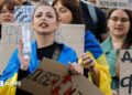 Ukrainians, including a young woman draped in a Ukrainian blue-and-yellow flag, hold placards reading 'Bring him back" during a protest against the dismissal of Defence Minister Mykhailo Fedorov in downtown Kyiv