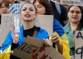 Ukrainians, including a young woman draped in a Ukrainian blue-and-yellow flag, hold placards reading 'Bring him back" during a protest against the dismissal of Defence Minister Mykhailo Fedorov in downtown Kyiv