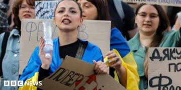 Ukrainians, including a young woman draped in a Ukrainian blue-and-yellow flag, hold placards reading 'Bring him back" during a protest against the dismissal of Defence Minister Mykhailo Fedorov in downtown Kyiv