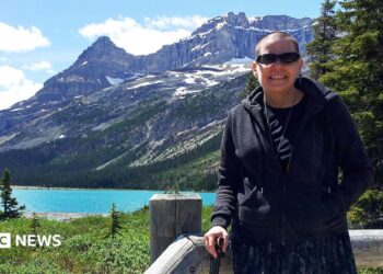 Cindy Baldassi is in the foreground wearing sunglasses and a dark hoody and navy blue dress. She is looking at the camera and behind her is a beautiful blue lake and towering mountain peaks with snow on top of them.
