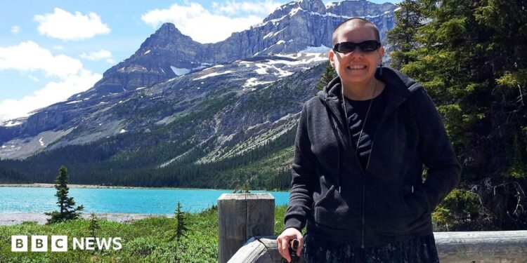 Cindy Baldassi is in the foreground wearing sunglasses and a dark hoody and navy blue dress. She is looking at the camera and behind her is a beautiful blue lake and towering mountain peaks with snow on top of them.