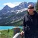 Cindy Baldassi is in the foreground wearing sunglasses and a dark hoody and navy blue dress. She is looking at the camera and behind her is a beautiful blue lake and towering mountain peaks with snow on top of them.