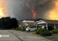 An image showing grey and orange smoke rising from behind a row of homes on a residential street in Summerland, British Columbia.