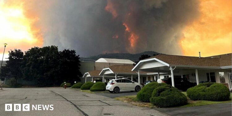 An image showing grey and orange smoke rising from behind a row of homes on a residential street in Summerland, British Columbia.