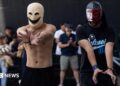 Two young men in masks hold their hands out.