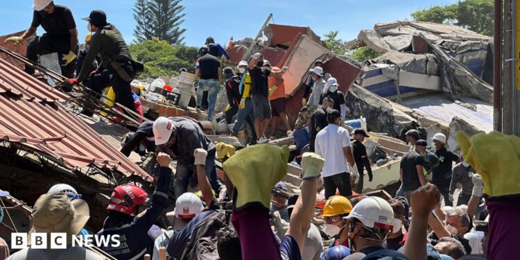 Alejandro Mahecha, he's a young person wearing a blue hard hat and a black t-shirt - he has medium length hair and has a serious expression