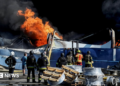 Wide shot showing Ukrainian firefighters in black and yellow uniforms observing flames rising from the debris of what appears to be a warehouse following a drone strike.