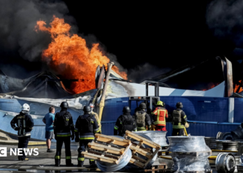 Wide shot showing Ukrainian firefighters in black and yellow uniforms observing flames rising from the debris of what appears to be a warehouse following a drone strike.