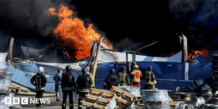 Wide shot showing Ukrainian firefighters in black and yellow uniforms observing flames rising from the debris of what appears to be a warehouse following a drone strike.