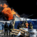 Wide shot showing Ukrainian firefighters in black and yellow uniforms observing flames rising from the debris of what appears to be a warehouse following a drone strike.