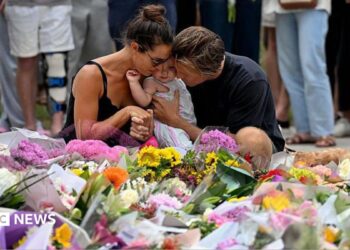 A woman with dark hair wearing a black strappy top and a man with dark blonde hair wearing a black shirt hug a young child as they kneel at a floral tribute to the Bondi Beach shooting victims at the Bondi Pavillion in Sydney on 15 December, 2025.