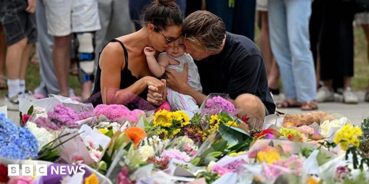 A woman with dark hair wearing a black strappy top and a man with dark blonde hair wearing a black shirt hug a young child as they kneel at a floral tribute to the Bondi Beach shooting victims at the Bondi Pavillion in Sydney on 15 December, 2025.
