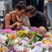 A woman with dark hair wearing a black strappy top and a man with dark blonde hair wearing a black shirt hug a young child as they kneel at a floral tribute to the Bondi Beach shooting victims at the Bondi Pavillion in Sydney on 15 December, 2025.