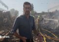 Dan Johnson wears a dark shirt and glasses, standing amongst rubble with smoke in the air and firefighers using a hose in the background