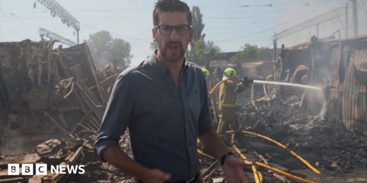 Dan Johnson wears a dark shirt and glasses, standing amongst rubble with smoke in the air and firefighers using a hose in the background