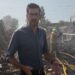 Dan Johnson wears a dark shirt and glasses, standing amongst rubble with smoke in the air and firefighers using a hose in the background