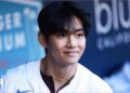 V smiles as he looks at a distance, wearing a baseball shirt