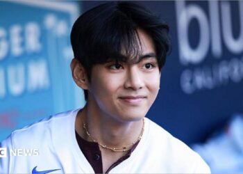 V smiles as he looks at a distance, wearing a baseball shirt