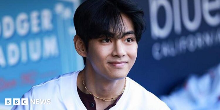 V smiles as he looks at a distance, wearing a baseball shirt