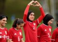 Five women in red football kit stand in a line. The one in the middle is holding her arms above her head and her hands form the shape of a heart.