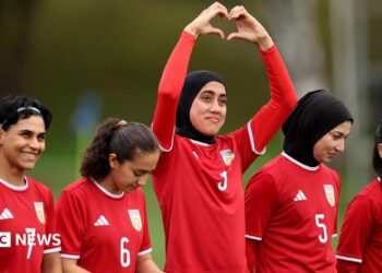 Five women in red football kit stand in a line. The one in the middle is holding her arms above her head and her hands form the shape of a heart.
