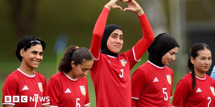 Five women in red football kit stand in a line. The one in the middle is holding her arms above her head and her hands form the shape of a heart.