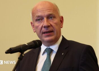 Berlin Mayor Kai Wegner speaks into a microphone on a pale yellow background wearing a dark suit and blue tie.