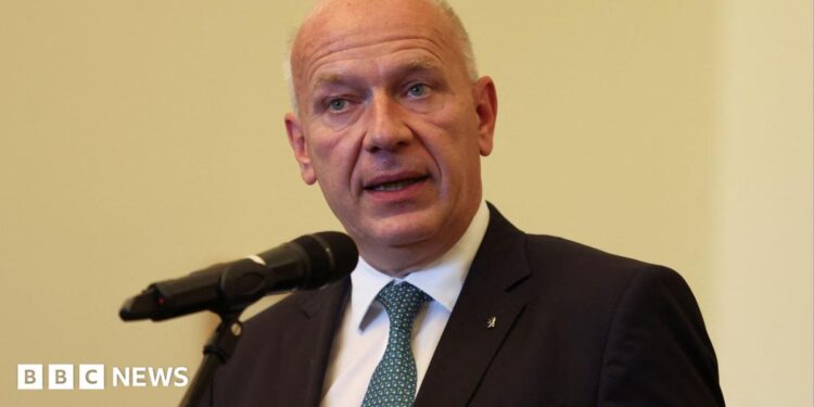 Berlin Mayor Kai Wegner speaks into a microphone on a pale yellow background wearing a dark suit and blue tie.