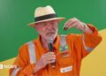 President Lula, wearing an orange jumpsuit with the logo of the state-run oil company Petrobras, hold up a small container filled with oil. He is wearing a cream-coloured Panama hat with a black ribbon around it. In his right hand, he is holding a microphone.