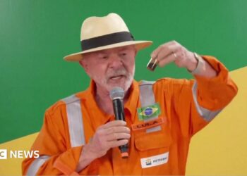 President Lula, wearing an orange jumpsuit with the logo of the state-run oil company Petrobras, hold up a small container filled with oil. He is wearing a cream-coloured Panama hat with a black ribbon around it. In his right hand, he is holding a microphone.