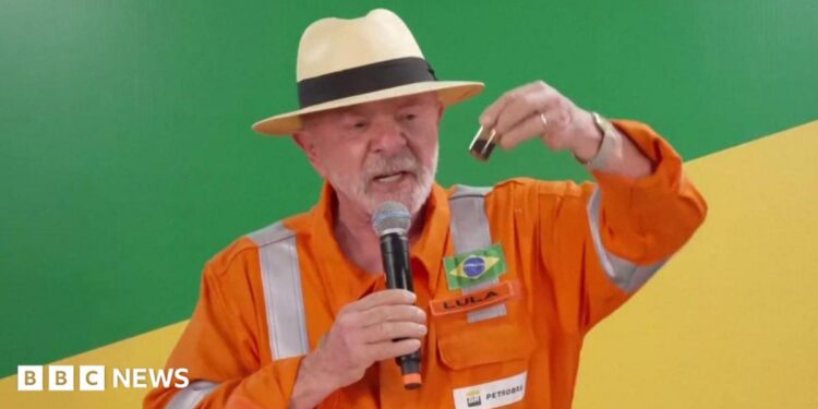 President Lula, wearing an orange jumpsuit with the logo of the state-run oil company Petrobras, hold up a small container filled with oil. He is wearing a cream-coloured Panama hat with a black ribbon around it. In his right hand, he is holding a microphone.
