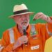 President Lula, wearing an orange jumpsuit with the logo of the state-run oil company Petrobras, hold up a small container filled with oil. He is wearing a cream-coloured Panama hat with a black ribbon around it. In his right hand, he is holding a microphone.