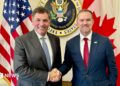 An image showing US trade representative Jamieson Greer (right) shaking hands with Canada-US trade minister Dominic LeBlanc to his left. Behind them are a US and Canada flag with the seal for the US trade representative in the middle. Both men are wearing suits and smiling at the camera.