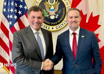 An image showing US trade representative Jamieson Greer (right) shaking hands with Canada-US trade minister Dominic LeBlanc to his left. Behind them are a US and Canada flag with the seal for the US trade representative in the middle. Both men are wearing suits and smiling at the camera.