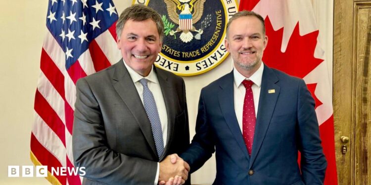 An image showing US trade representative Jamieson Greer (right) shaking hands with Canada-US trade minister Dominic LeBlanc to his left. Behind them are a US and Canada flag with the seal for the US trade representative in the middle. Both men are wearing suits and smiling at the camera.