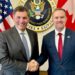 An image showing US trade representative Jamieson Greer (right) shaking hands with Canada-US trade minister Dominic LeBlanc to his left. Behind them are a US and Canada flag with the seal for the US trade representative in the middle. Both men are wearing suits and smiling at the camera.