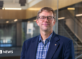 An image showing Prof Guikema. He has short dirty blonde hair, and round glasses with thin black frames. He is slightly smiling. He is wearing a navy blue smart coat with a checkered blue button-up shirt underneath. Behind him is a backdrop of an academic building, showing glass panels and stairs that are blurred.