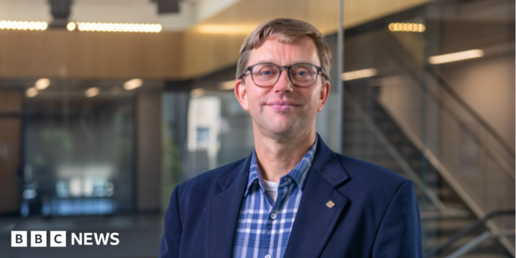 An image showing Prof Guikema. He has short dirty blonde hair, and round glasses with thin black frames. He is slightly smiling. He is wearing a navy blue smart coat with a checkered blue button-up shirt underneath. Behind him is a backdrop of an academic building, showing glass panels and stairs that are blurred.