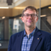 An image showing Prof Guikema. He has short dirty blonde hair, and round glasses with thin black frames. He is slightly smiling. He is wearing a navy blue smart coat with a checkered blue button-up shirt underneath. Behind him is a backdrop of an academic building, showing glass panels and stairs that are blurred.