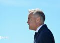 Mark Carney stands in profile, facing to the left, against a clear light blue sky. He wears a dark blue suit and tie.