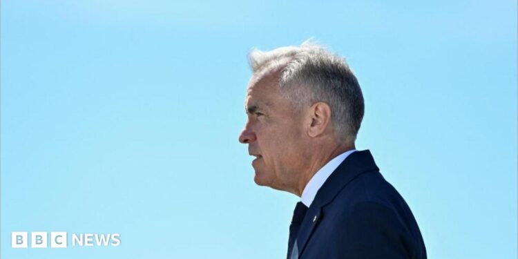 Mark Carney stands in profile, facing to the left, against a clear light blue sky. He wears a dark blue suit and tie.